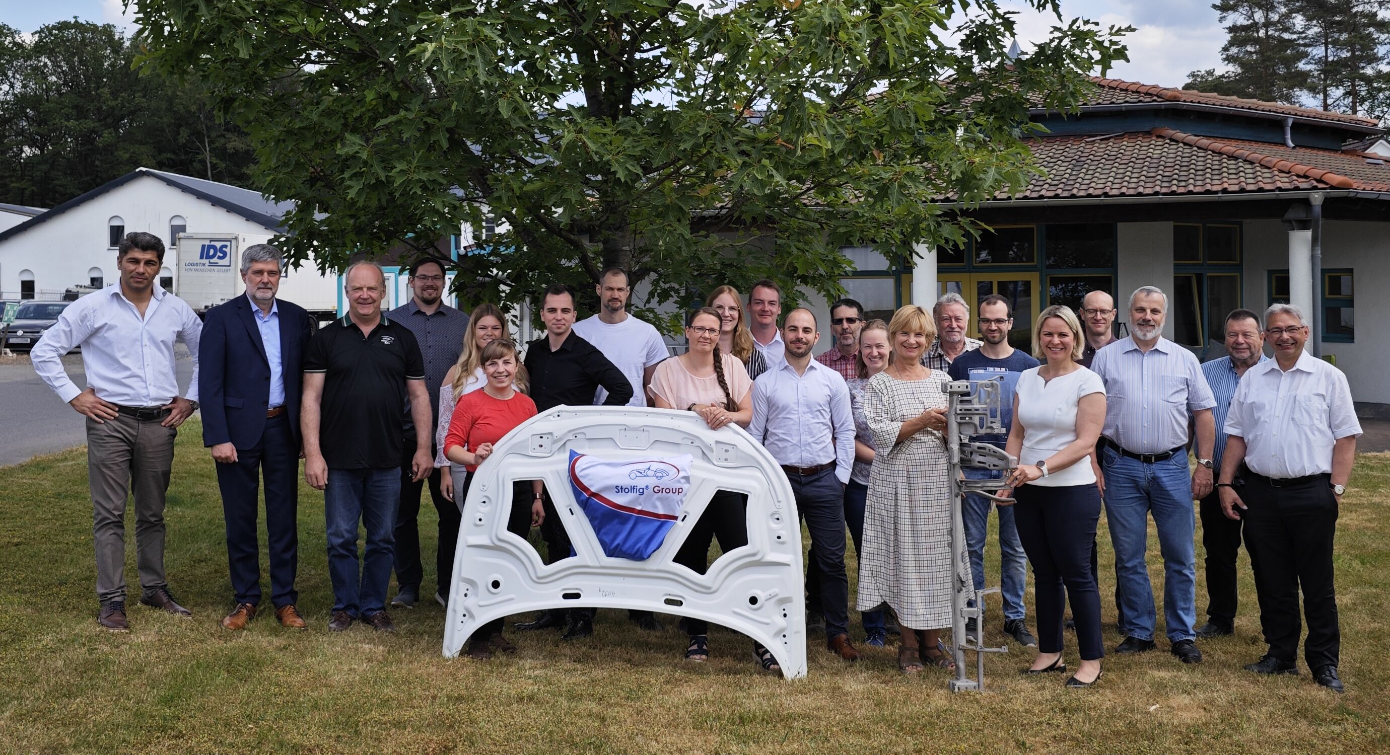 20 Vertreter der CLEAN-Mag Projektpartnerunternehmen posieren auf einer Grünfläche mit Leichtbaukomponenten aus Magnesium in der Hand.