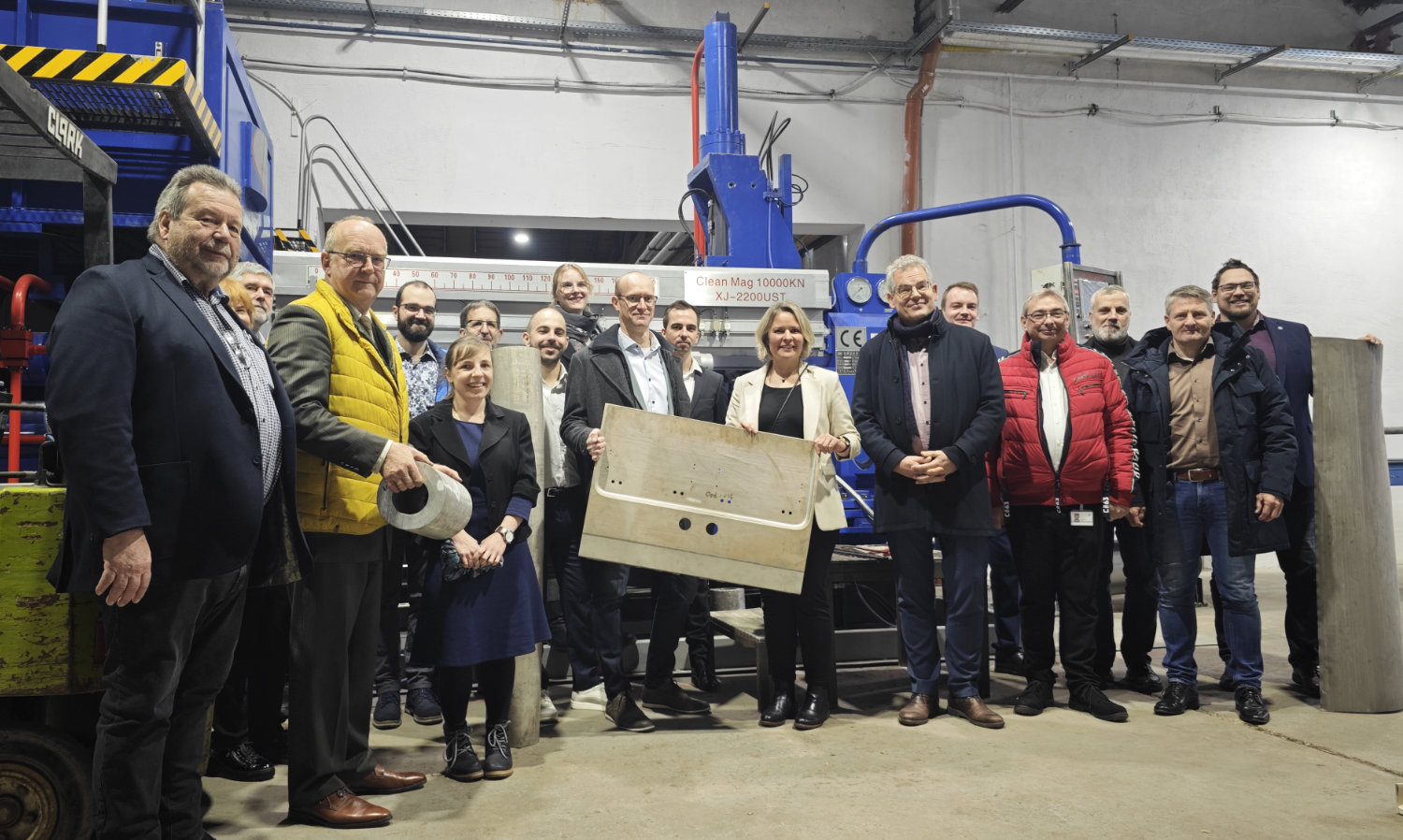Gruppenfoto der CLEAN-Mag-Projektpartner beim Abschlusstreffen in einer der Produktionshallen der STOLFIG LEICHTBAU KOMPETENZ ZENTRUM GMBH: Mehrere Teilnehmende stehen vor einer großen blauen Strangpressanlage. In der Mitte werden ein warmumgeformtes Bauteil aus dem Transportsketor sowie ein hohles Magnesiumbillet als Beispiele der Projektergebnisse präsentiert.